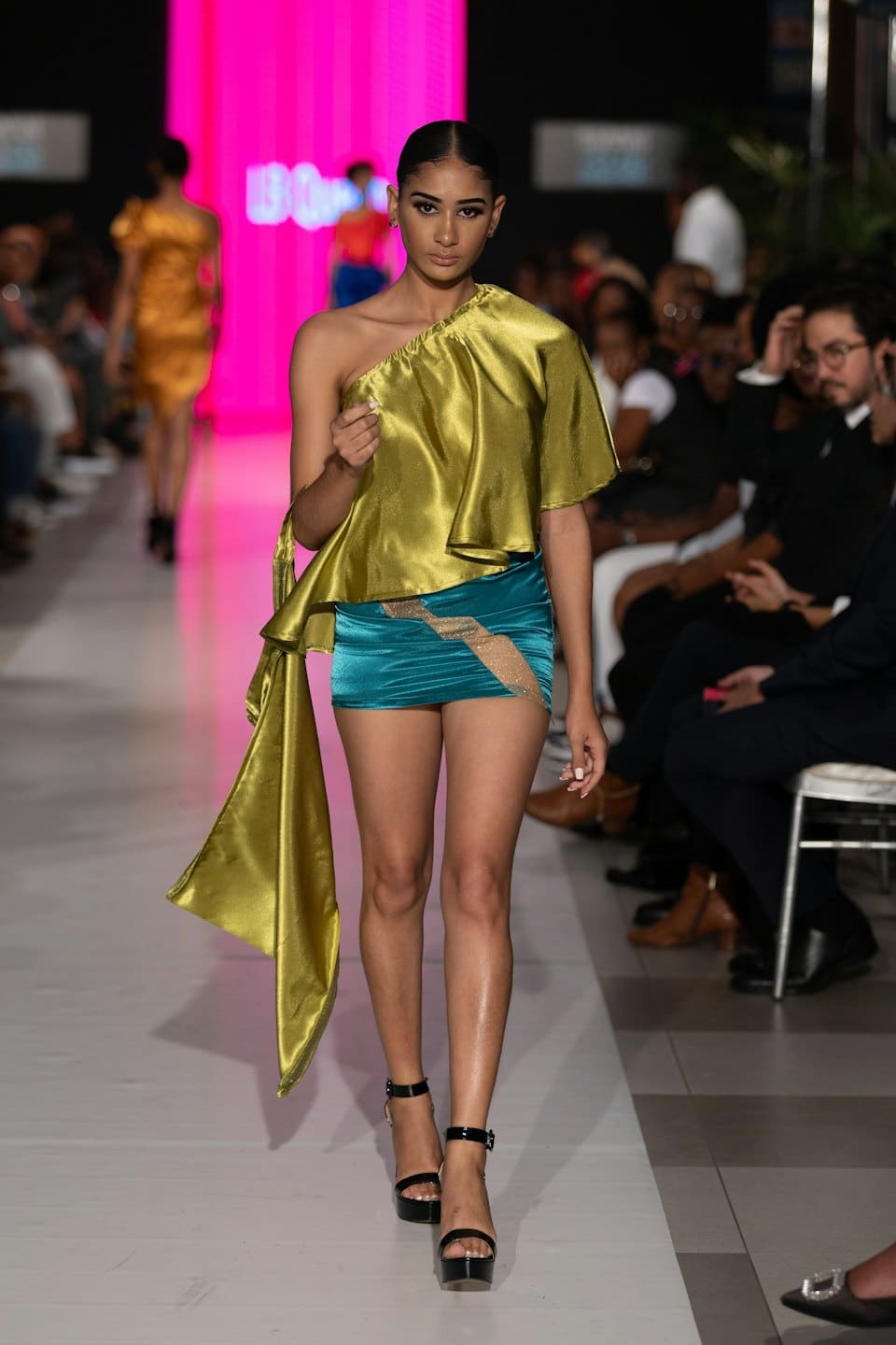 Model walking on a runway wearing a chartreuse top and teal skirt.