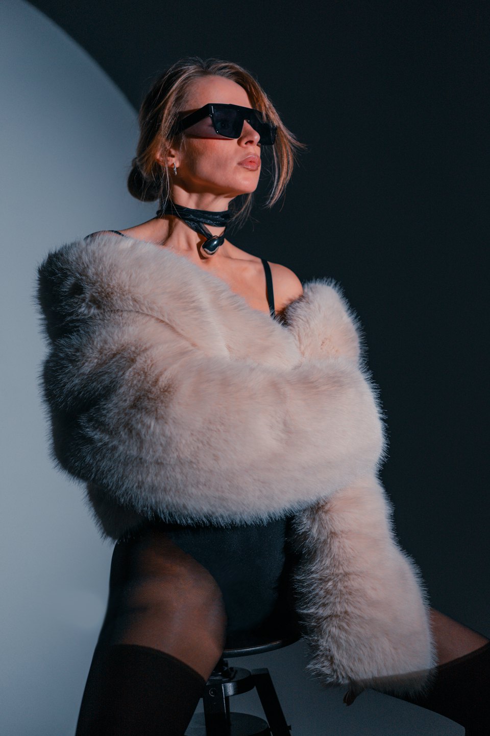 Woman in a beige fur coat sits on a stool.